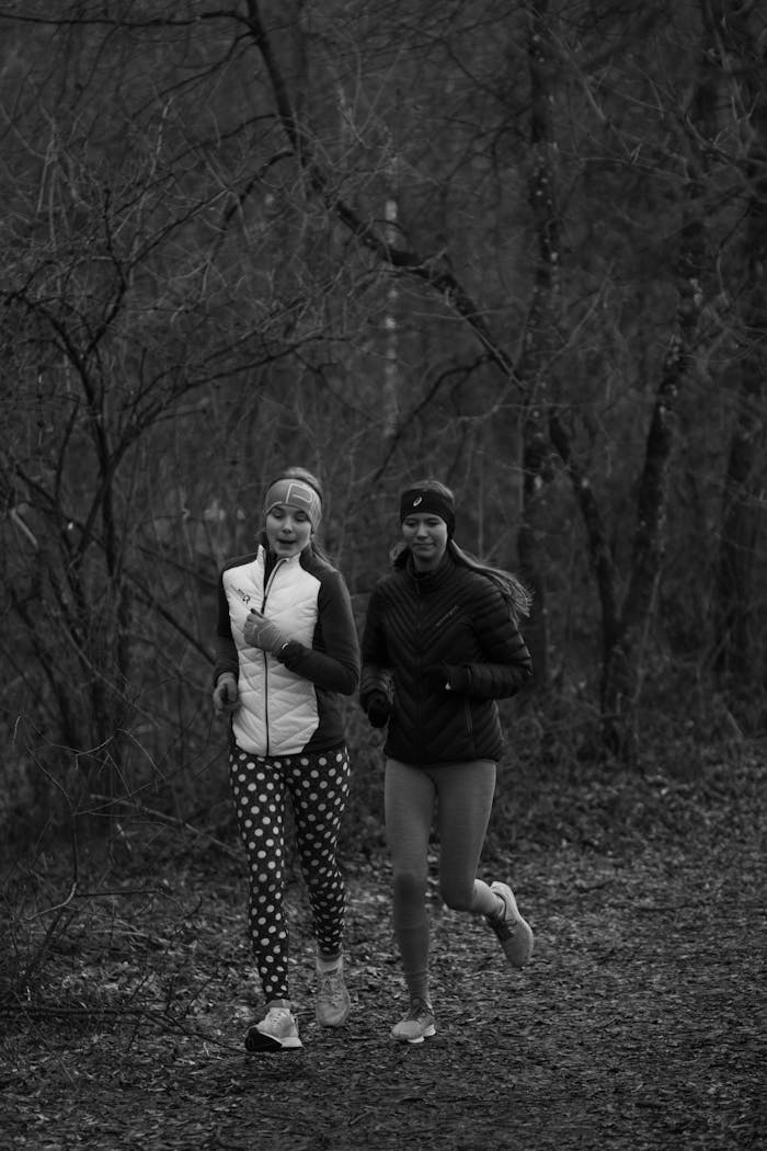 Two women running through a forest path during the day wearing sportswear, captured in black and white.
